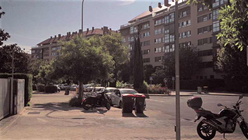 Quiet residential street featuring parked vehicles and motorcycle, revealing urban neighborhood scenery with buildings, trees, and sunny perspectives