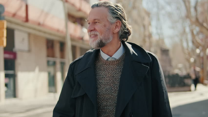 Happy senior man with grey hair and beard smiles while walking in a city. He wears a sweater and overcoat on a sunny day.