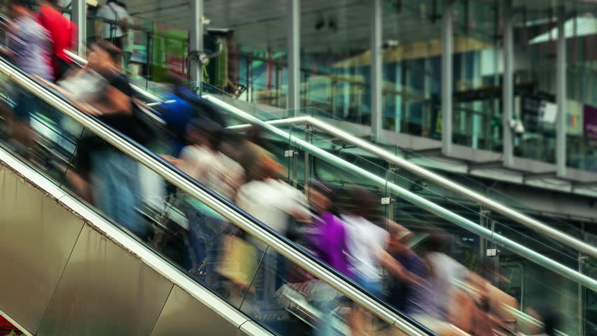 Large crowd of people moves upwards on an escalator in a modern commercial or transportation hub. The long exposure shot creates a dynamic motion blur effect, depicting the fast pace and constant