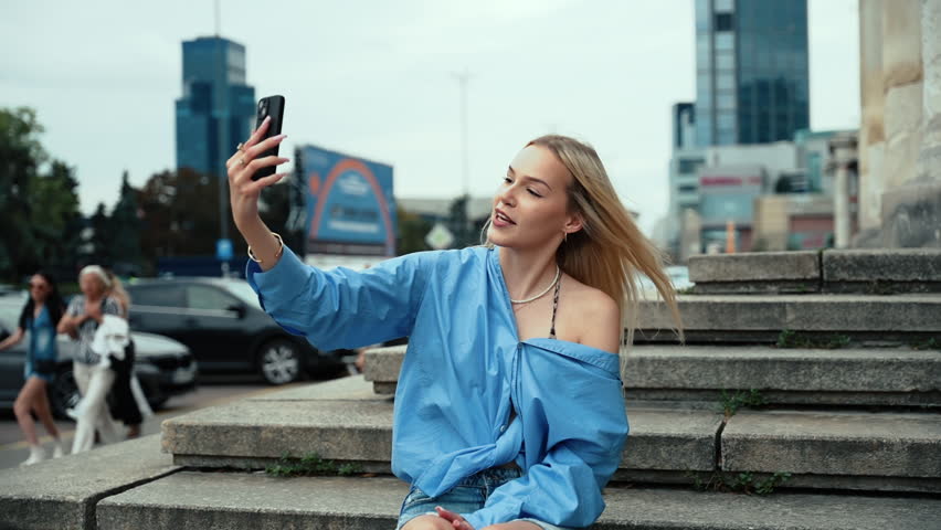 Young blonde woman in a blue summer shirt and denim shorts sitting on stone steps, happily video calling with joyful emotions.