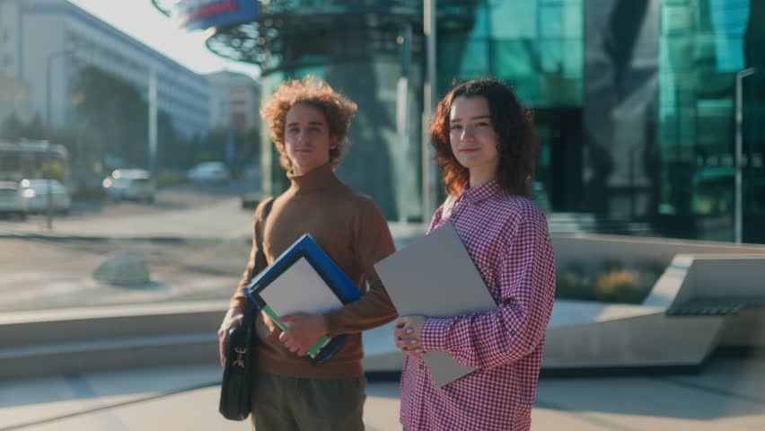 Two youthful students, a young woman and man, stand together outside a modern university campus with glass walls, holding folders and enjoying academic lifestyle.