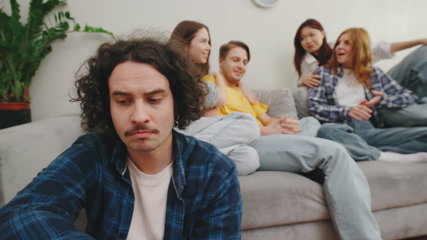 A young man sits on the floor beside a sofa, refusing to engage with two couples who try to talk to him, creating a moment of emotional distance in a cozy apartment