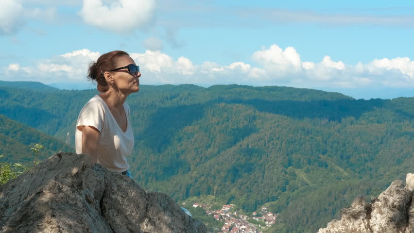 Woman relaxing on mountain top enjoying sunny day. Woman sits on a rock, soaking in the stunning view of a town nestled in a valley. Surrounded by lush trees and majestic mountains