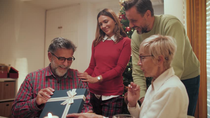 Multigenerational family members gathering near christmas tree, warmly presenting wrapped gift to delighted senior grandfather during festive holiday moment