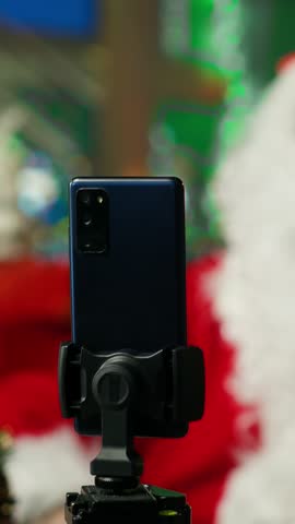 Vertical video Santa Claus using smartphone on tripod to film vlog opening letters from kids. Older person wearing Christmas costume recording video with phone selfie camera, reading letters from
