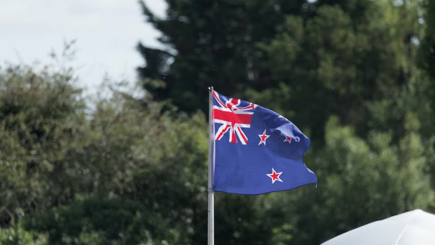 New Zealand flag waving on flagpole outdoors, surrounded by green trees in natural daylight.