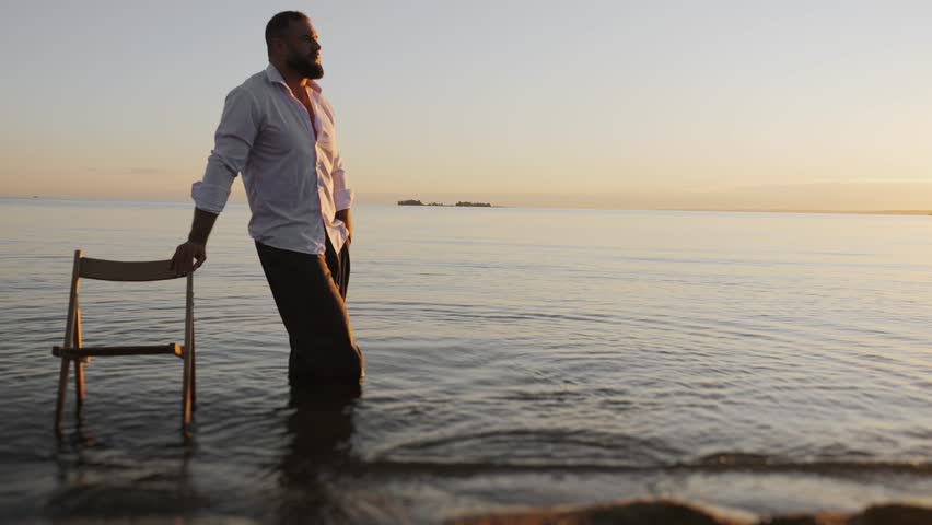 A brutal man in pants and a white shirt stands in the water and holds on to a chair at sunset in the evening on the seashore.