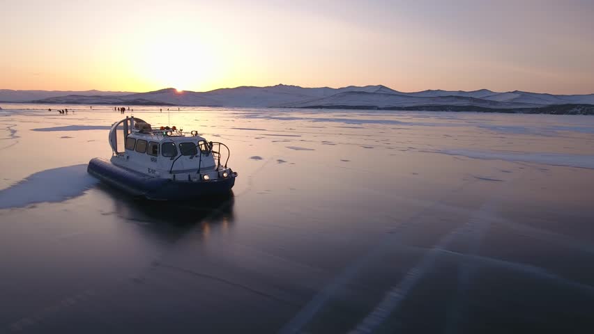 Aerial air cushion vehicle moves across mirror-like ice of Lake Baikal during magnificent orange sunset. Tourist expedition to Siberia