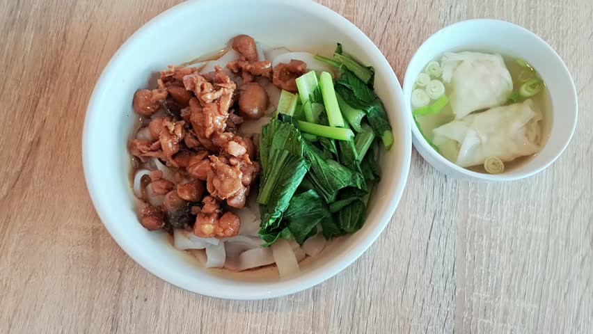 A flat lay view of the boewl of Kwetiau noodlewith vegetables and savory chicken; and a bowl of dumplings soup.