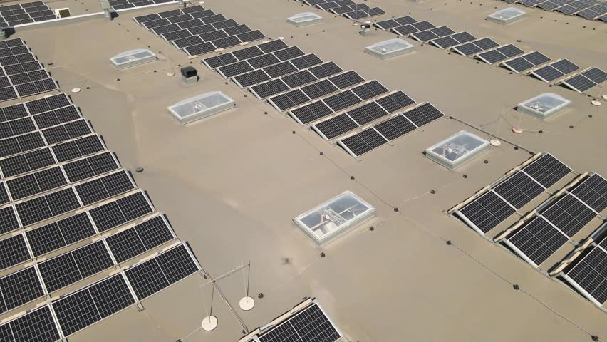 Aerial drone video panning over a rooftop that has hundreds of renewable energy creating solar panels that are dark and black colored and are shining under the sunny weather. Ventilation systems also.