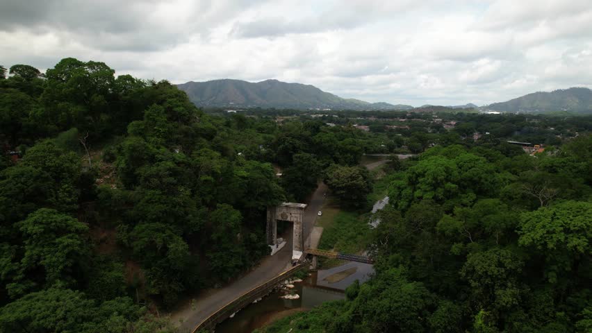 breathtaking drone shot pan right, reveal iconic "Puerta del Llano" monument in Venezuela, historic stone tunnel and bridge over Guárico River. Lush natural landscape with winding roads and mountains,