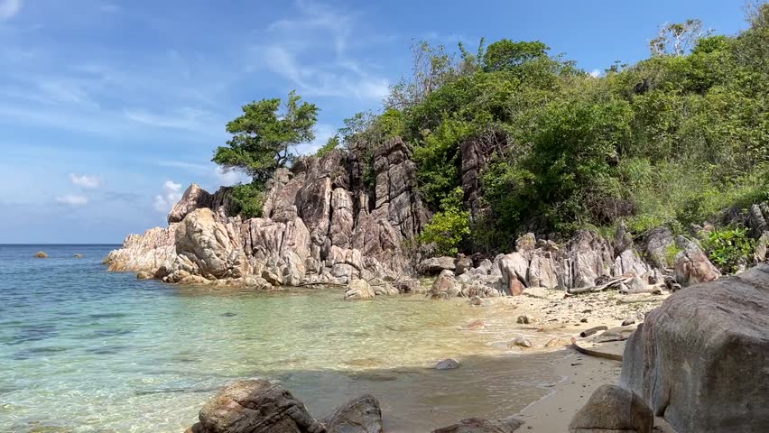 Captures serene rocky coastline near Lamai, Ko Samui, where jagged cliffs meet turquoise waters. Lush greenery under a clear sky creates a tranquil and inviting landscape.