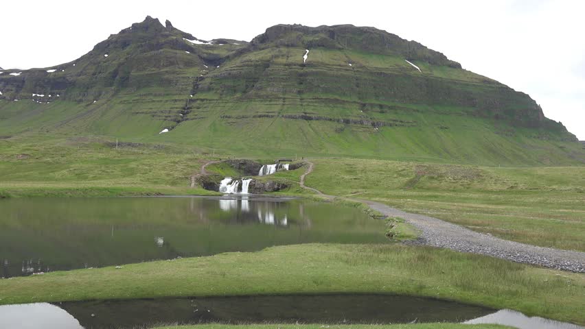 Picturesque view of Kirkjufellsfoss waterfall in the foreground with the dramatic peak of Kirkjufell rising just beyond. The scene highlights the harmony between rushing water, rugged volcanic rock