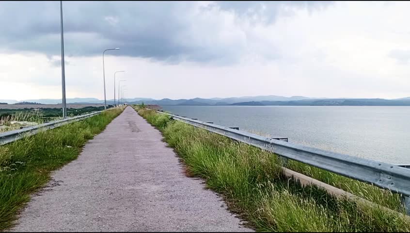 Dark clouds gather above a peaceful lake lined with a winding path. Streetlights stand sentinel among swaying grasses. A gravel shore edges the still water, with hills vanishing into the gloomy sky