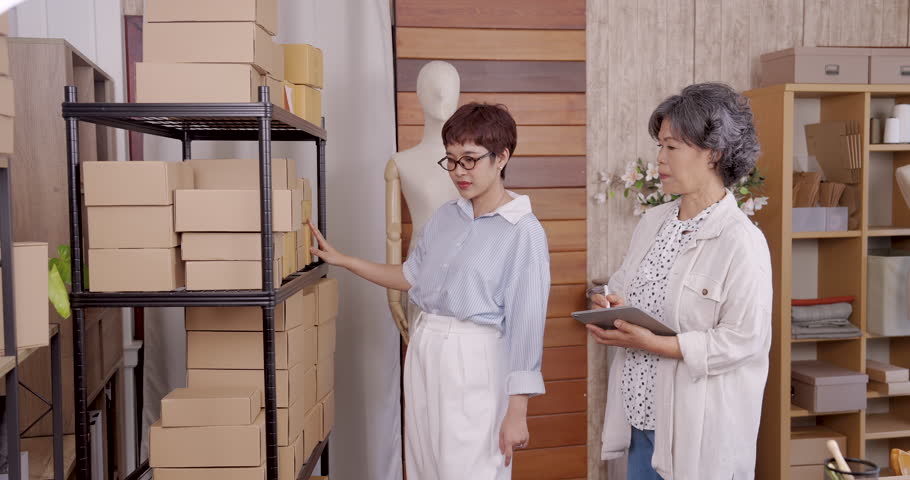 Asian adult female daughter pointing shelf while asian senior female mother reviewing tablet during stocktake inside family boutique atelier storage rack full boxes teamwork small business clothing