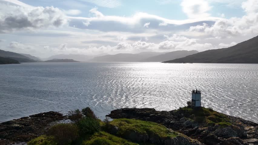 Seabirds fly over sunlit loch, lighthouse, and dramatic clouds in the Scottish Highlands panorama.