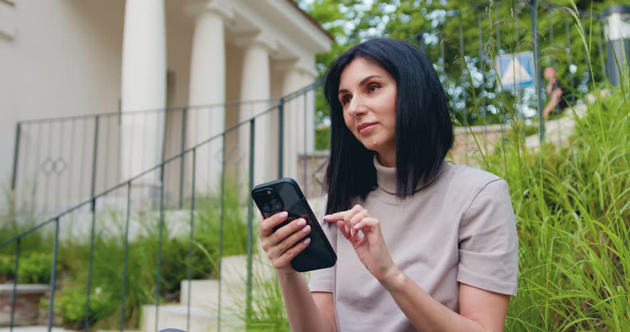Pretty young brunette woman holding smartphone, scrolling social media texting browsing online against the background of modern old building. Outdoors
