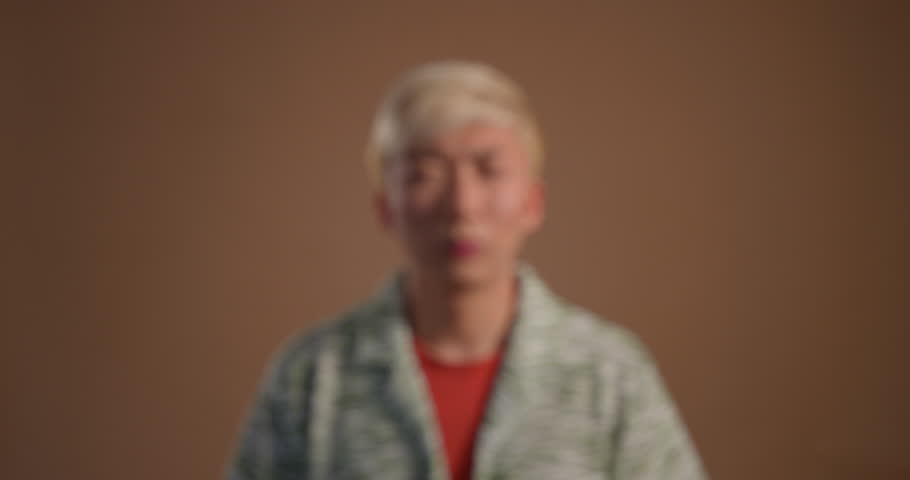 Video clip of a young adult male looking for and putting on his glasses in the studio. The clip begins blurred and comes into focus as he puts on his glasses. He has bleached hair and is wearing casua