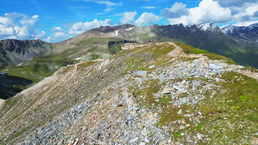 Aerial view of the majestic mountains near Glocknerstraße, where rugged terrain meets lush greenery under a blue sky dotted with fluffy clouds, Bruck an der Großglocknerstraße, Salzburg, Austria.