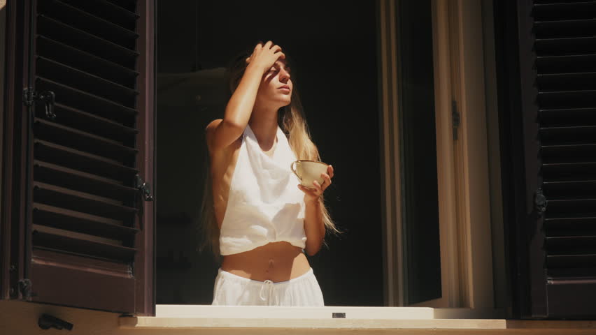 Woman in white clothing enjoying morning sunlight while drinking coffee at an open window