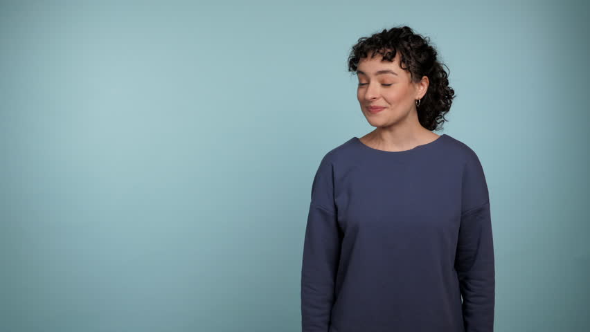 Smiling young curly woman in blue sweater pointing finger to the side workspace promo area mock up and presenting product. Portrait positive beautiful female standing on isolated light blue background