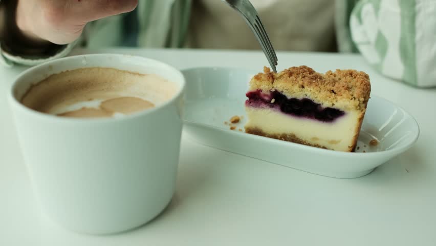 Close-up of a hand holding a fork with a slice of blueberry cheesecake next to a cup of cappuccino in cozy cafe. Young woman eating cake in a cafe. Horizontal 4k footage