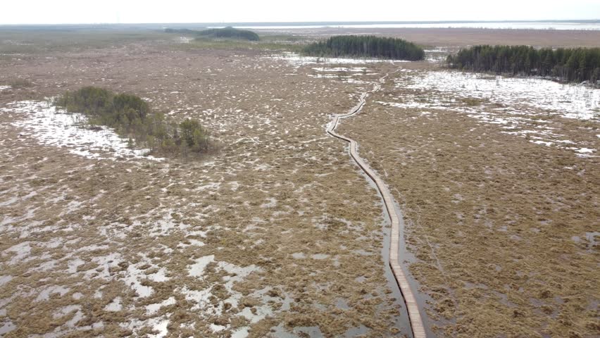 A wide aerial view of a vast swamp landscape, featuring a long, winding boardwalk stretching into the distance amidst patches of snow and marshland