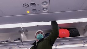 Masked traveler placing backpack in overhead while boarding flight, Man loads backpack into overhead bin on airplane, safety signs lit, travel, winter clothes, preparing for flight, cabin scene - Powered by Shutterstock - Get 15% off with code: PIKWIZARD15