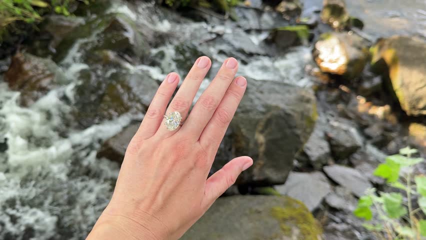 A landscape video of a sparkling oval diamond engagement ring or wedding band on platinum band displayed above river rocks in Scandinavian sunlight in Sweden.