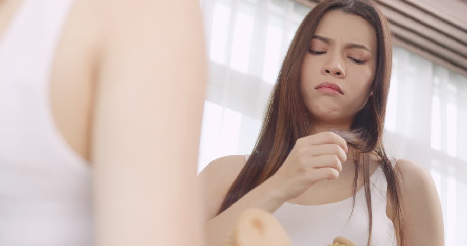 A young woman is worried about her hair health while looking in the mirror at home.