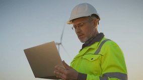 Male engineer in helmet and reflective jacket holding laptop. Worker standing outdoors near wind turbine. Looking into distance. Technician monitoring renewable energy project on site. - Powered by Shutterstock - Get 15% off with code: PIKWIZARD15