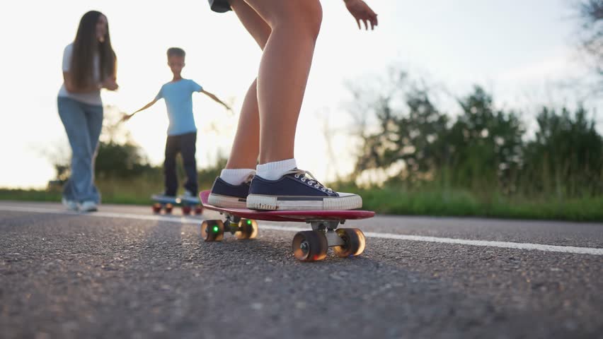 A girl on a skateboard moves swiftly down the road, her sneakers firmly on the skateboard. Behind, a boy skateboards on the road while talking to the girl on the road.