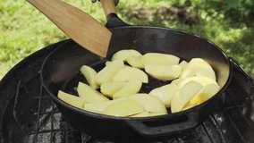 Close-up of outside on the grill coals, potatoes cut into slices are being fried in a cast iron pan. The cook is stirring the potatoes in the pan with a wooden spatula. Slow motion video 100fp. - Powered by Shutterstock - Get 15% off with code: PIKWIZARD15