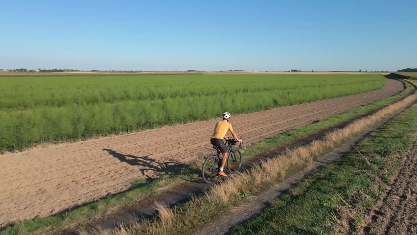 Cyclist rides along rural dirt road between green fields under blue sky. Man in helmet and cycling uniform training on gravel bike on countryside road. Concept of adventure travel and active lifestyle