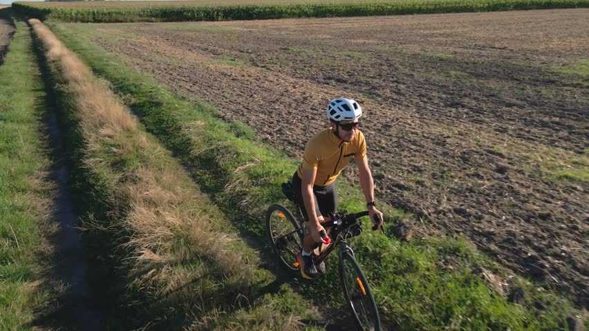 Cyclist rides along rural dirt road between green fields under blue sky. Man in helmet and cycling uniform training on gravel bike on countryside road. Concept of adventure travel and active lifestyle