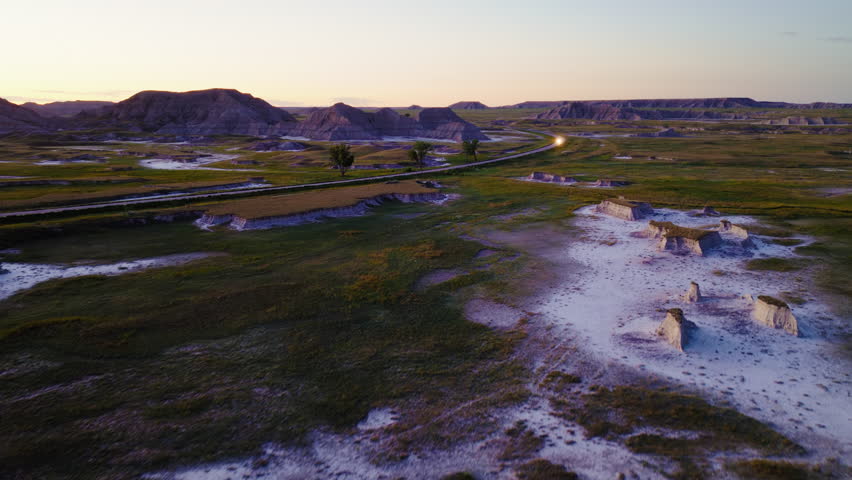 Sunset aerial views capture a scenic road cutting across rugged Badlands, with golden light highlighting dramatic terrain, deep shadows, and the raw beauty of the landscape