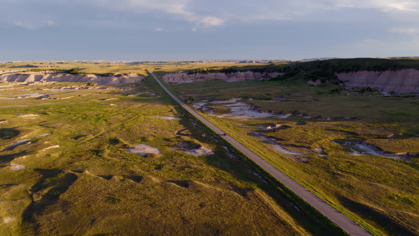 A serene sunset glows over the Badlands as a lonely road winds through rugged peaks, with warm colors and dramatic shadows creating a peaceful yet powerful landscape scene