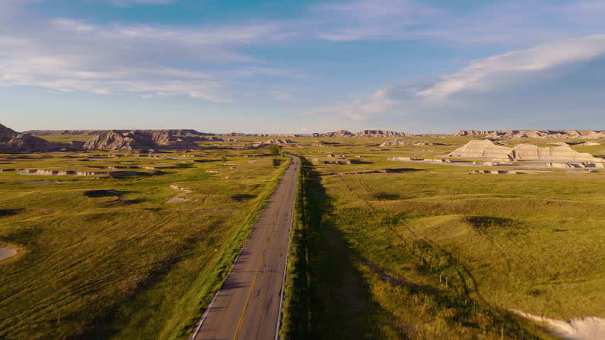 A winding road snakes through rugged Badlands terrain, captured by drone at sunset as warm light highlights dramatic formations and the remote beauty of the landscape unfolds