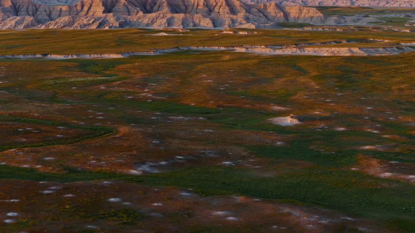 Sunset glow illuminates harsh Badlands ridges in aerial flight, casting golden light and deep shadows across rugged terrain for a breathtaking view of this dramatic landscape