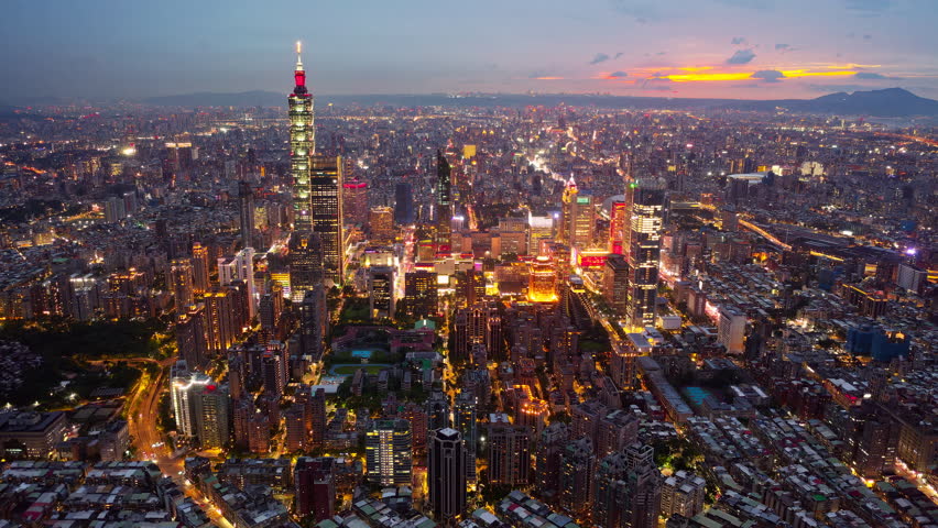 Aerial hyperlapse above XinYi District in Downtown Taipei, vibrant capital of Taiwan, where 101 Tower stands out amid modern skyscrapers and city lights dazzle in twilight under dramatic sunset sky