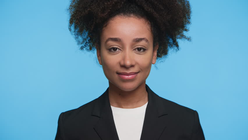 Smiling young woman in black blazer holding her hand to her ear, expressing curiosity and excitement while listening against blue background.
