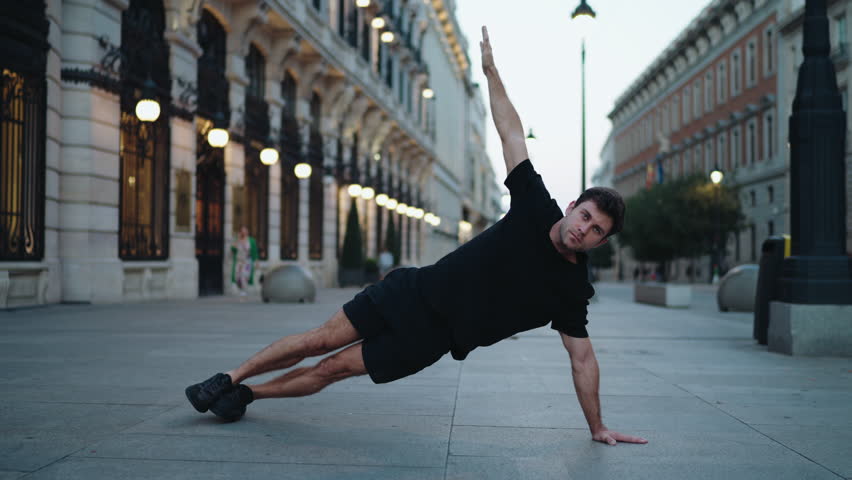 Man in black sportswear holding side plank on urban street, raising one arm for balance. Concept of core strength, stability, and dedication in early morning workout routine