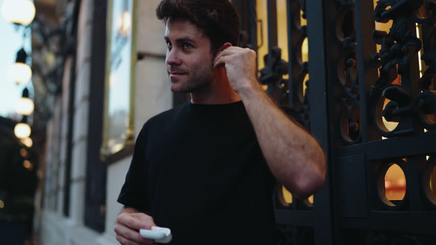 Smiling man holding case and wearing wireless earbuds before workout in city. Represents positive lifestyle, preparation, fitness motivation, and use of audio technology outdoors
