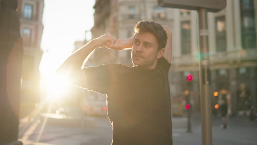 Athletic man stretching arms overhead in city at sunrise, wearing sportswear. Symbol of preparation, discipline, and active healthy lifestyle with vitality