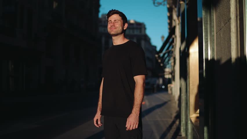 Man standing in sunlight on urban street with eyes closed, smiling peacefully. Symbol of relaxation, positivity, and balance in modern lifestyle