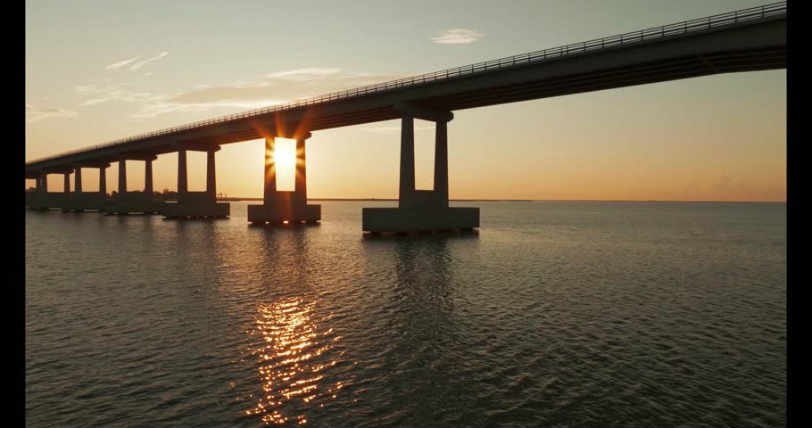 Flying Drone View of Road Bridge Over Coastal Water With Bright Sun at Sunset