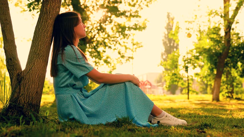 Young smiling girl, sits under tree in city park. Travel concept, young woman, nature. Girl student resting enjoying landscape of park in summer. Meditation young woman on green grass. Beautiful girl