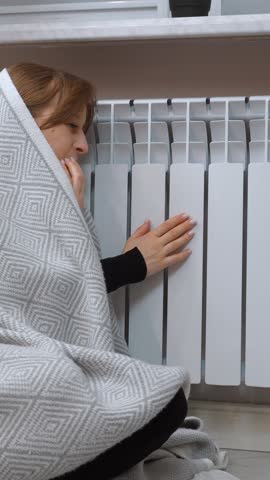 Freezing woman covered in blanket warming up near radiator at home, trying to get warm during cold winter season, concept of high price of heating, energy crisis and expensive bills vertical video