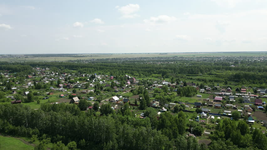 Aerial view panning across a typical russian village with colorful houses, surrounded by green trees and fields, under a bright summer sky