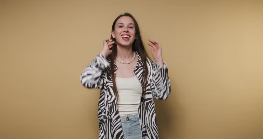 A woman joyfully raises her arms in celebration, wearing a trendy zebra-patterned jacket and a white top. The vibrant backdrop enhances the cheerful mood of the moment.